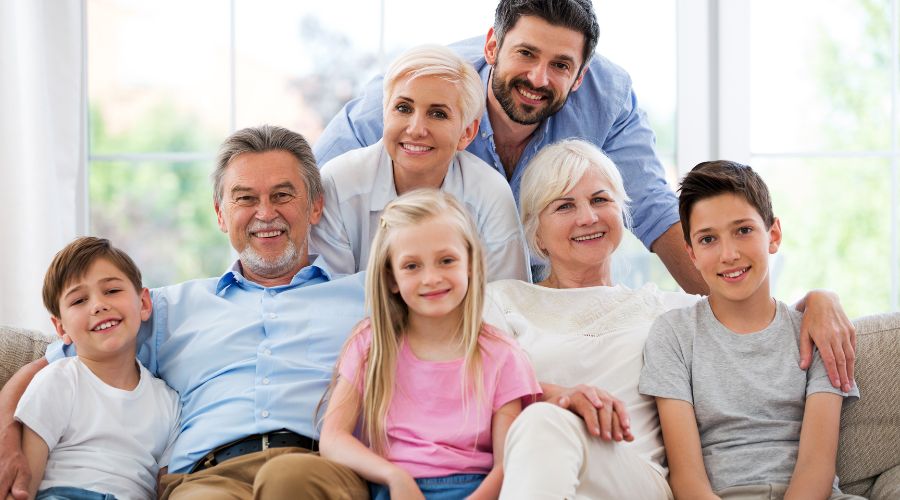 foto di famiglia sorridente, mamma e papà in piedi dietro al divano, nonno e nonna seduti accanto a una nipotina e due nipotini